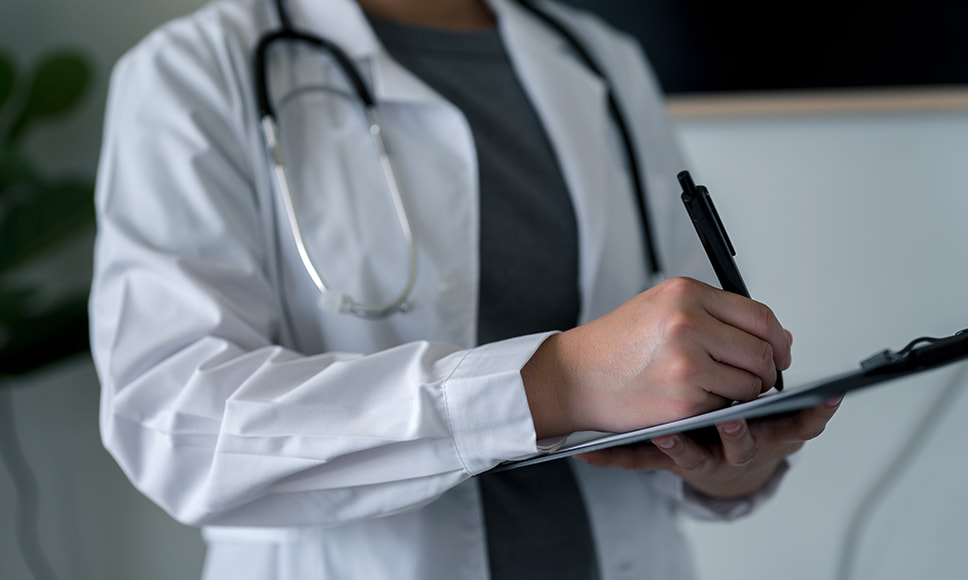 Medico in camice bianco con stetoscopio che scrive su una cartella clinica.
